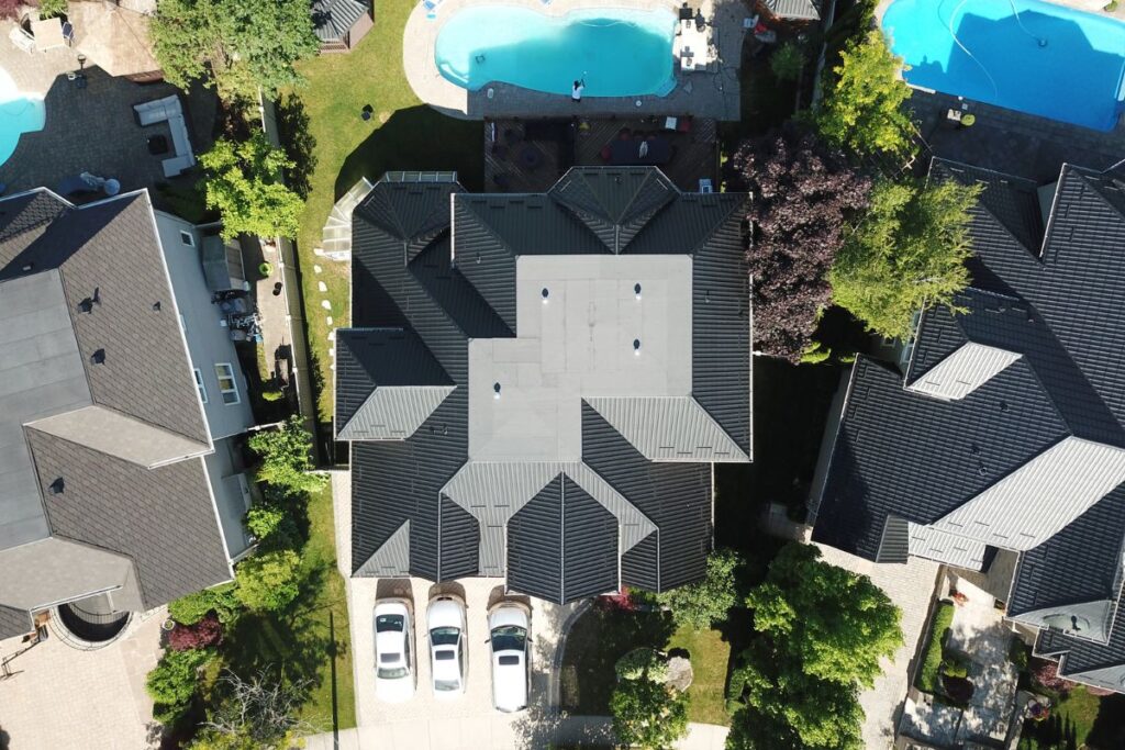 Aerial view of a large home with a sleek metal tile roof, a well-maintained driveway with several cars, and a spacious backyard featuring a pool.