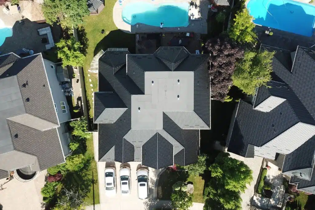 Aerial view of a large home with a sleek metal tile roof, a well-maintained driveway with several cars, and a spacious backyard featuring a pool.