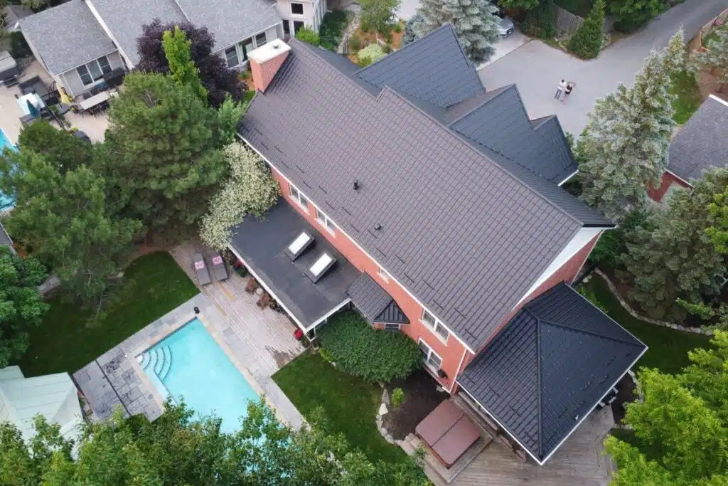 Aerial view of a spacious home with a sleek black metal roof, pool, and lush green trees in the backyard.