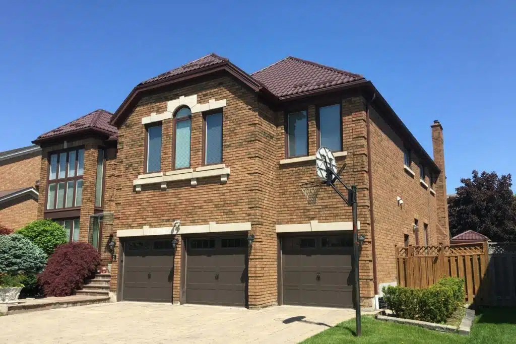 Grand brick home with a new brown metal roof, featuring a basketball hoop and lush greenery in the front yard.