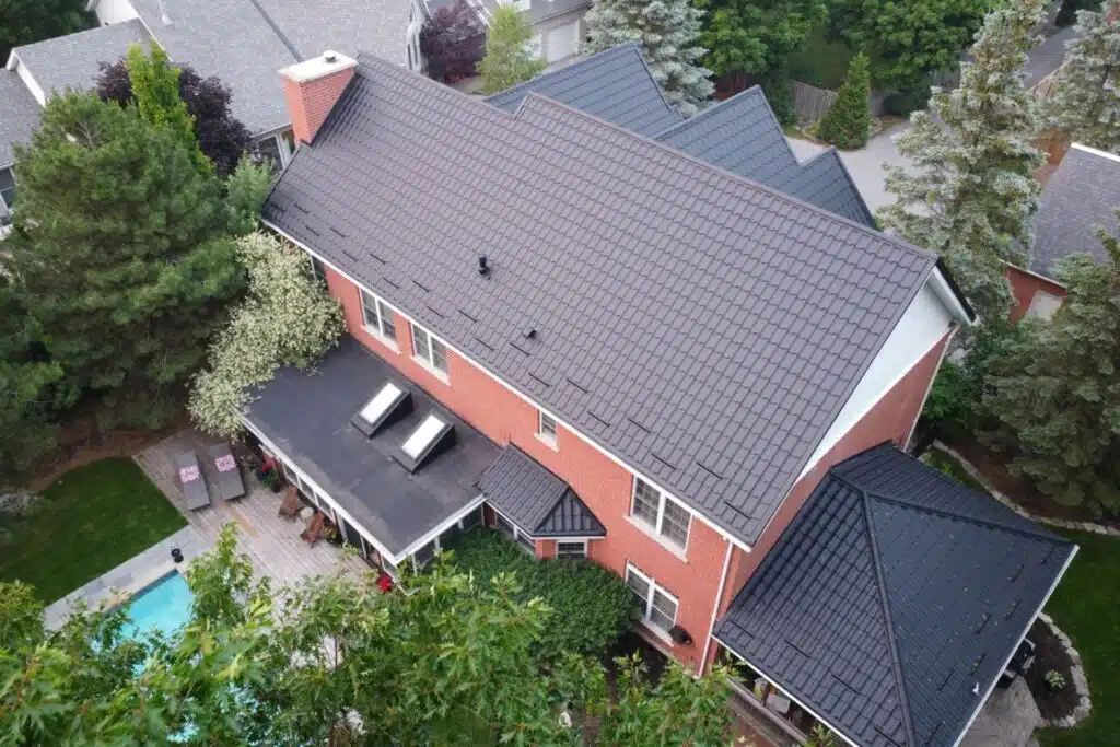 Aerial view of a brick home with a sleek black metal roof, a shaded patio area, and a pool surrounded by greenery.