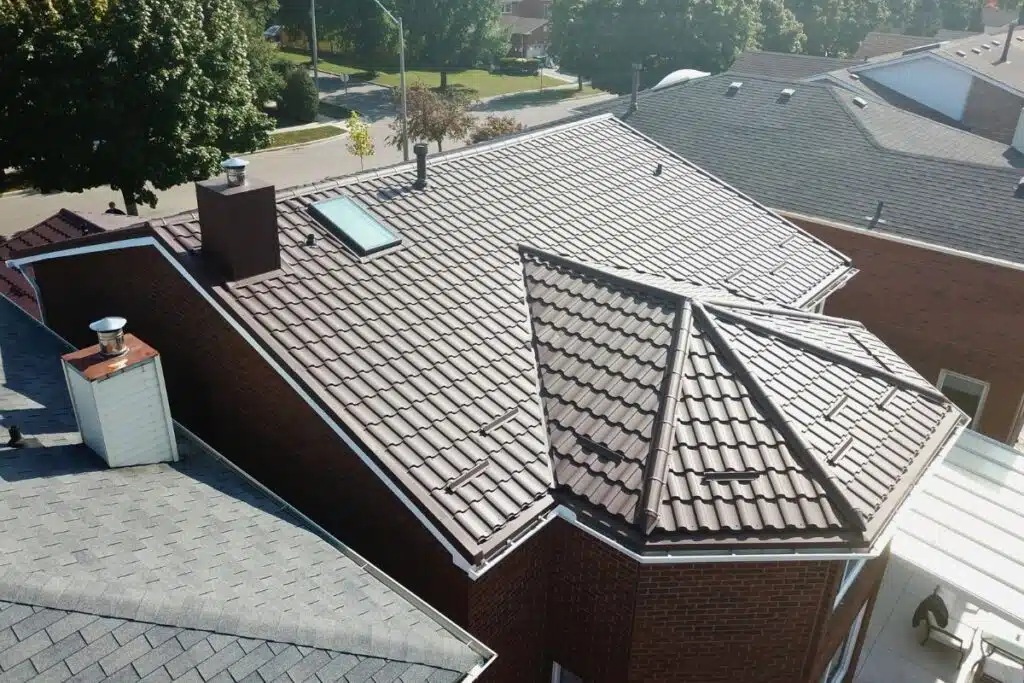 View of a brown metal roof installation with a skylight and chimney, contrasting against a nearby shingle roof.