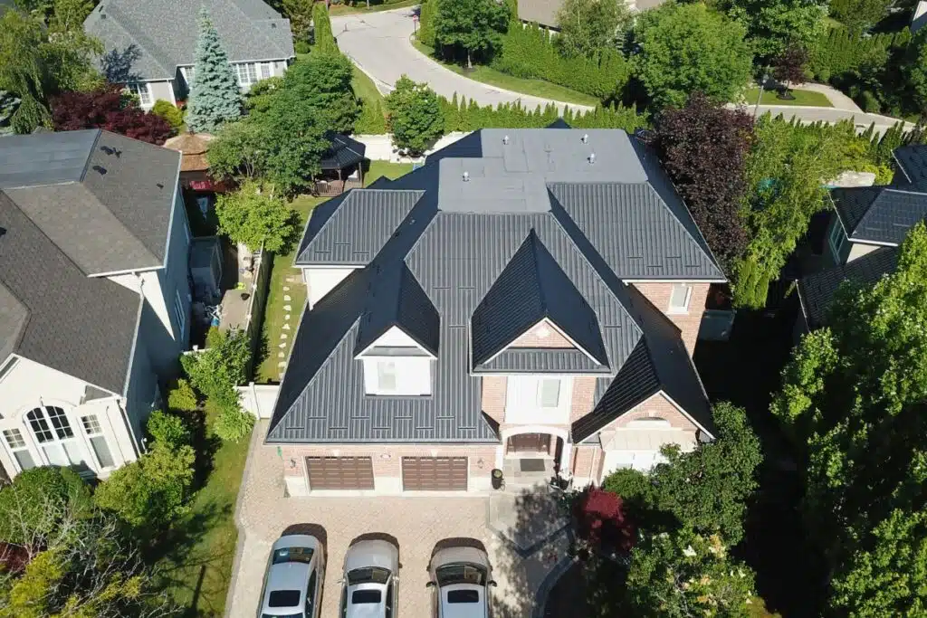 Beautiful aerial view of a large home with a sleek metal tile roof, surrounded by greenery, with a driveway full of parked cars.