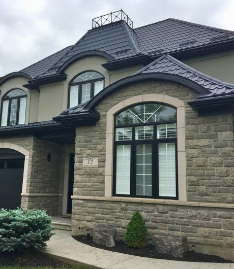 Metal Roofing Services in Burlington Front view of a house with a stone facade and dark blue metal roof, showcasing large windows and neatly manicured landscaping.