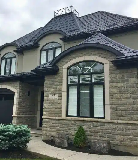 Front view of a house with a stone facade and dark blue metal roof, showcasing large windows and neatly manicured landscaping.