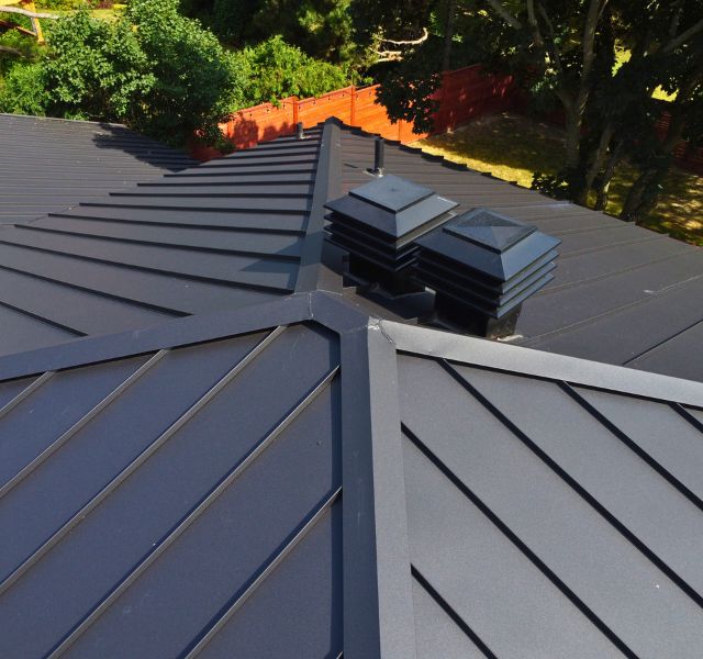 Modern Black Metal Roof with Vents – Durable Roofing Solution Close-up view of a black metal roof with distinct ridges and two roof vents.