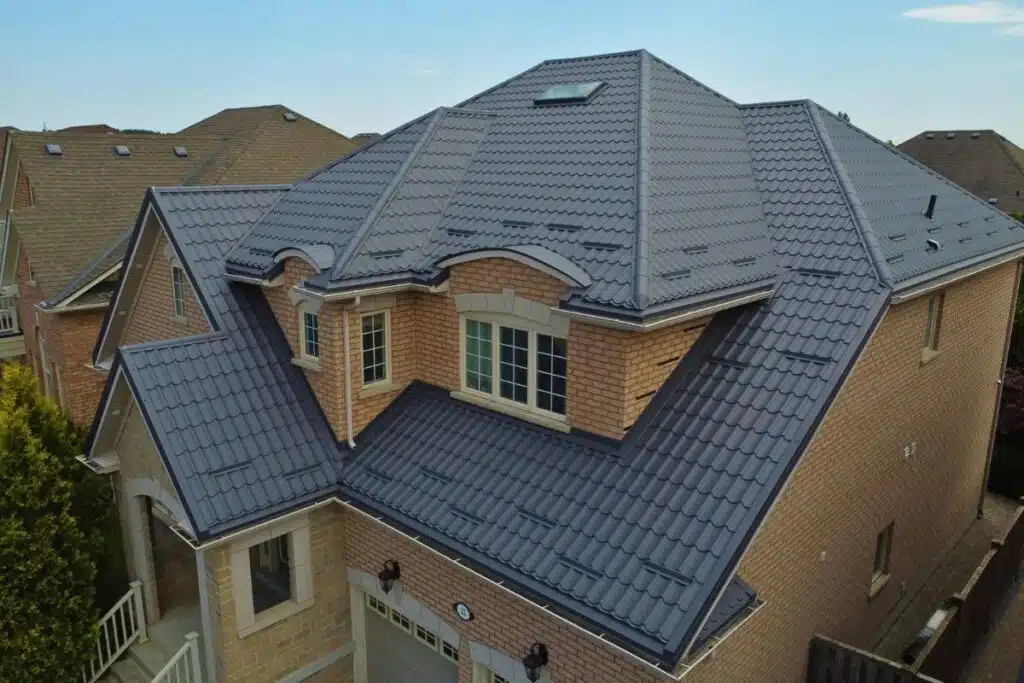 Large beige brick house with a dark grey Monterrey metal roof, featuring multiple dormer windows, a skylight, and a double garage.