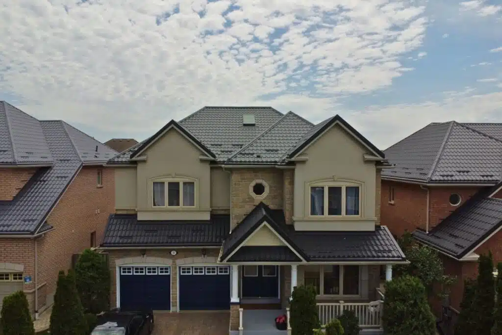 Two-story house with beige stucco, stone accents, and a dark grey Monterrey metal roof, featuring a double garage and covered front porch.