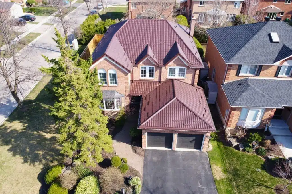 Two-story red brick house with a brown Monterrey metal roof, large windows, and a double garage, surrounded by a landscaped front yard.