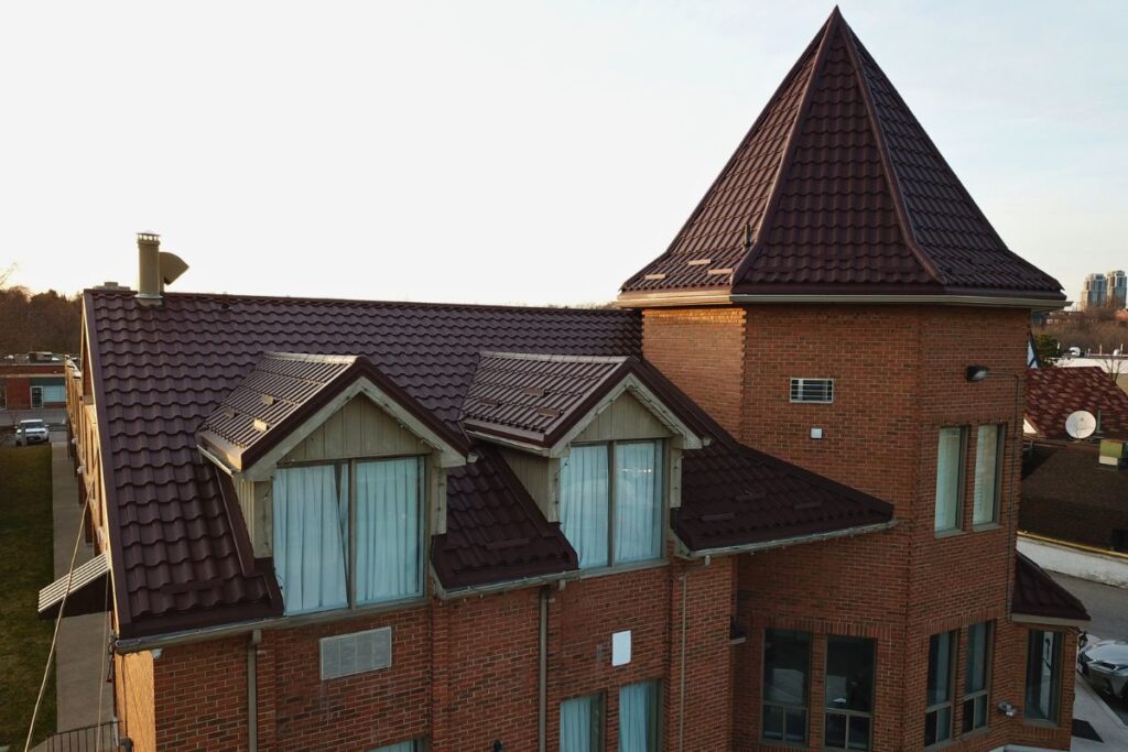 Red brick house with a brown Adamante metal roof featuring a tall turret and large windows, illuminated by the setting sun.