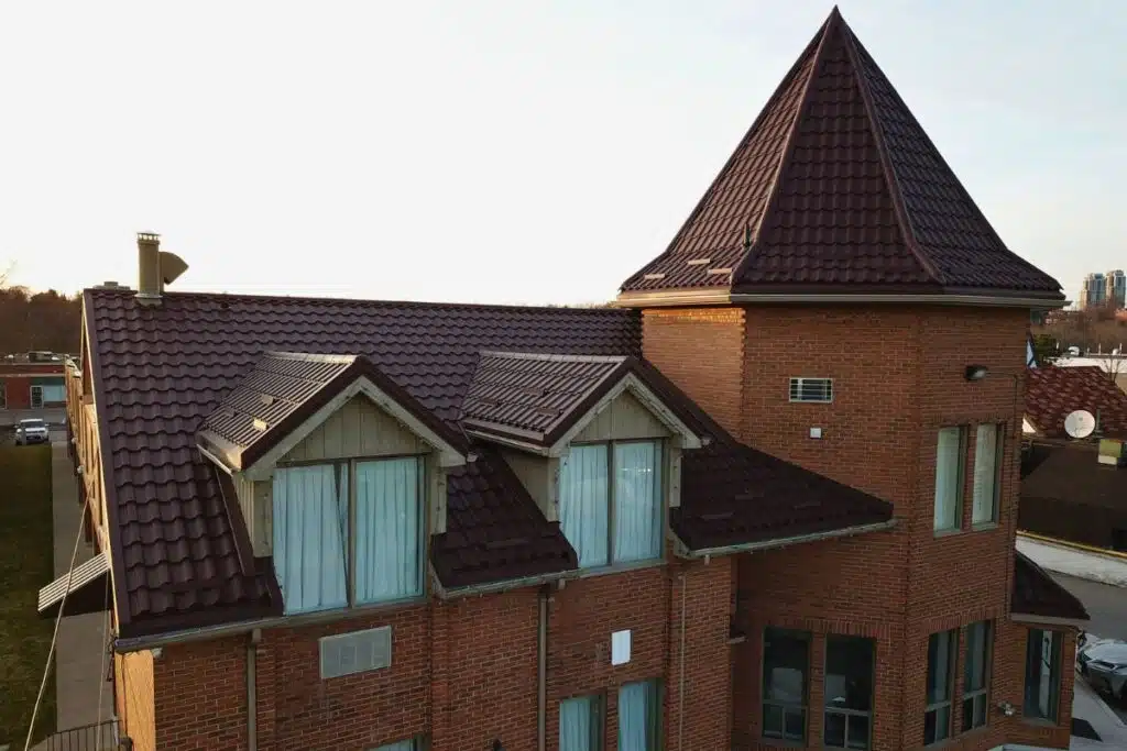 Elegant Adamante Metal Roof with Turret Design Red brick house with a brown Adamante metal roof featuring a tall turret and large windows, illuminated by the setting sun.