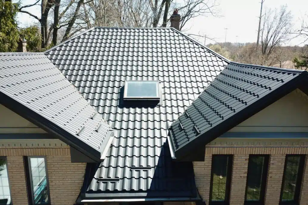 Adamante Metal Roof with Skylight – Modern and Durable Design Brick house with a dark grey Adamante metal roof, featuring a skylight in the center and surrounded by trees in the background.