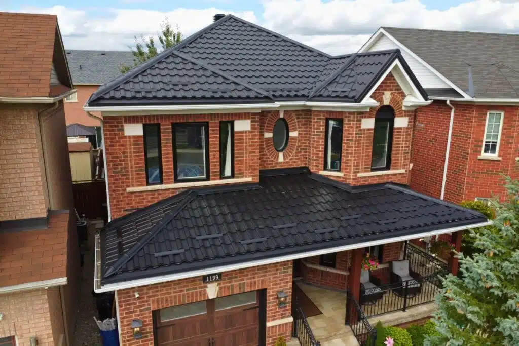 Stylish Adamante Metal Roof for Homes in Toronto Two-story red brick house with a dark grey Adamante metal roof, featuring large black-framed windows and a cozy front porch with seating.