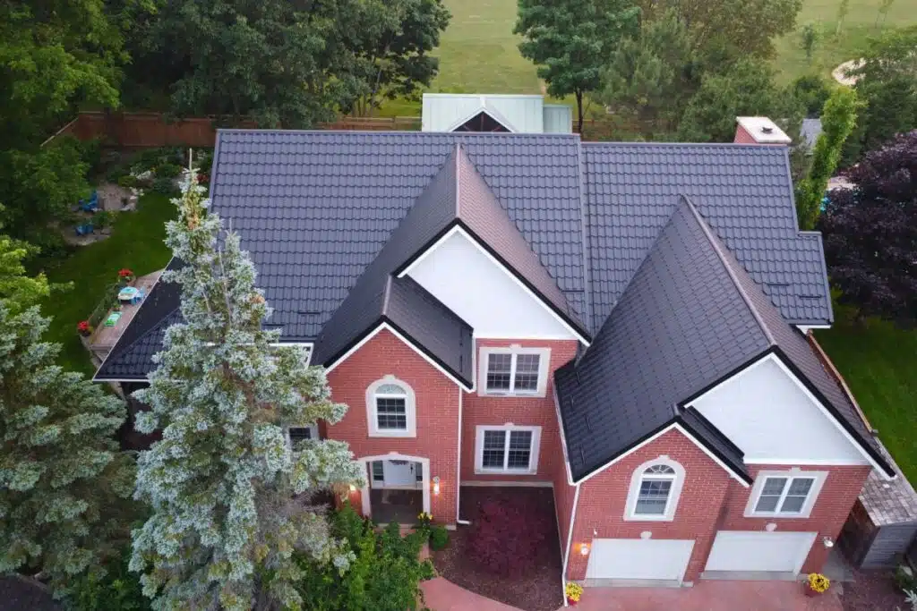 Adamante Metal Roof – Durable and Elegant Roof for Your Home Large brick house with white accents and a dark grey Adamante metal roof, surrounded by trees and a spacious green backyard.