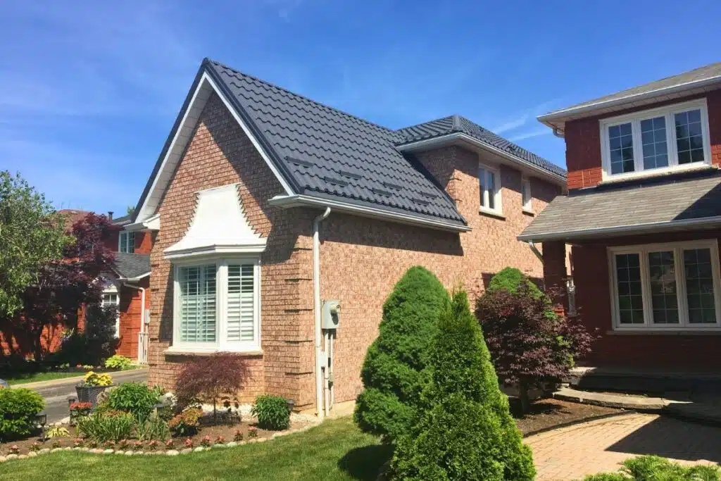 Adamante Metal Roofing – Modern Roof for Elegant Homes in Toronto Brick house with a dark grey Adamante metal roof, surrounded by a well-maintained garden and blue sky in the background.