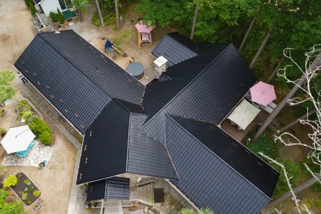 Elegant Black Metal Roof Installation in Tiny Township – Aerial View Aerial view of a large home in Tiny Township with a striking black metal roof, multiple gables, and surrounding greenery, showcasing its intricate layout and outdoor seating areas.