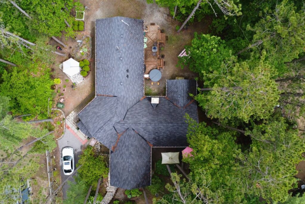 A top-down view of a home with a dark gray shingle roof, surrounded by lush greenery. The backyard features a stone pathway leading to a wooden deck with seating.
