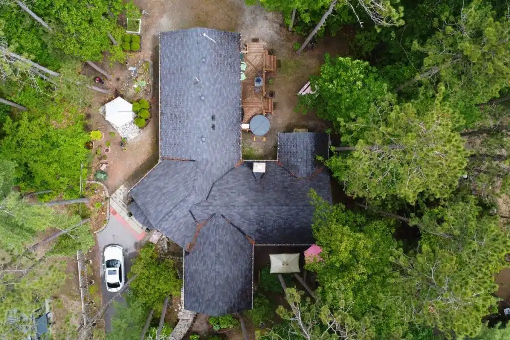 Dark Gray Shingle Roof with Lush Greenery and Stone Pathway A top-down view of a home with a dark gray shingle roof, surrounded by lush greenery. The backyard features a stone pathway leading to a wooden deck with seating.