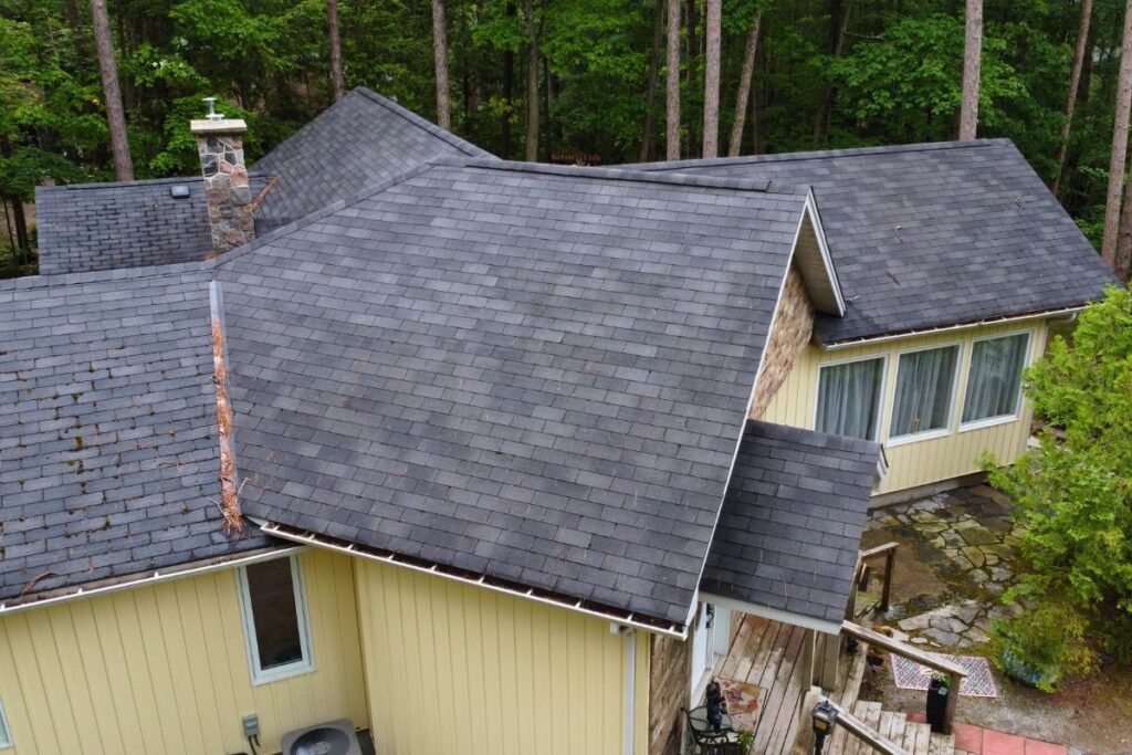 Aerial view of a sloping roof covered with dark gray shingles. Leaves and moss buildup along the edge. Below, a yellow wall with large windows and part of a stone pathway.