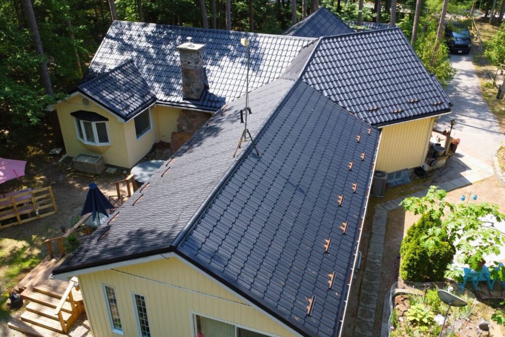 Aerial view of a large house with a metal roof featuring dark-colored tiles, a stone chimney, and a bright yellow wall. The surrounding environment includes trees and a paved pathway.