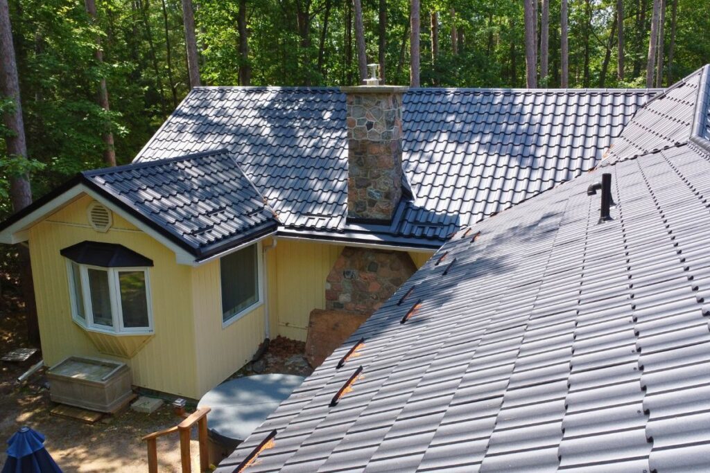 Aerial view of a roof with dark-colored metal tiles and a stone chimney in the center, surrounded by lush trees and part of a yellow building with a bay window.