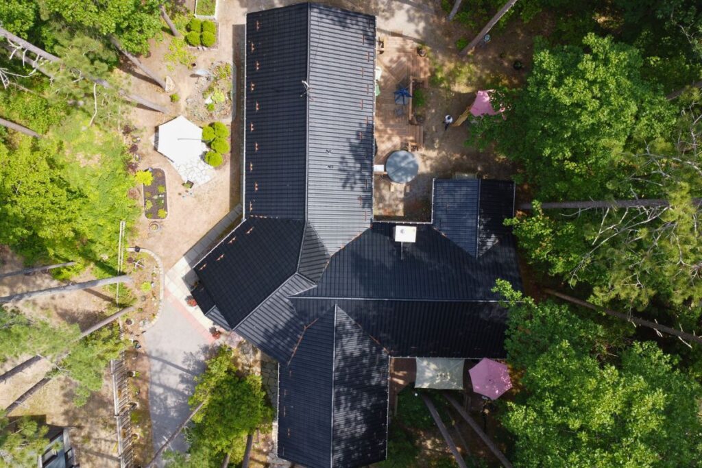 Aerial view of a large house with intersecting black metal roof slopes surrounded by lush green trees and a well-maintained outdoor area with garden and patio furniture.