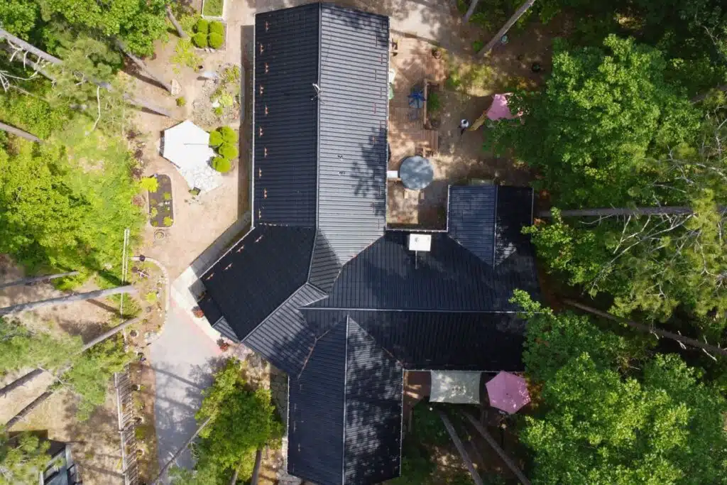 Large Black Metal Roof with Intersecting Slopes and Outdoor Area Aerial view of a large house with intersecting black metal roof slopes surrounded by lush green trees and a well-maintained outdoor area with garden and patio furniture.