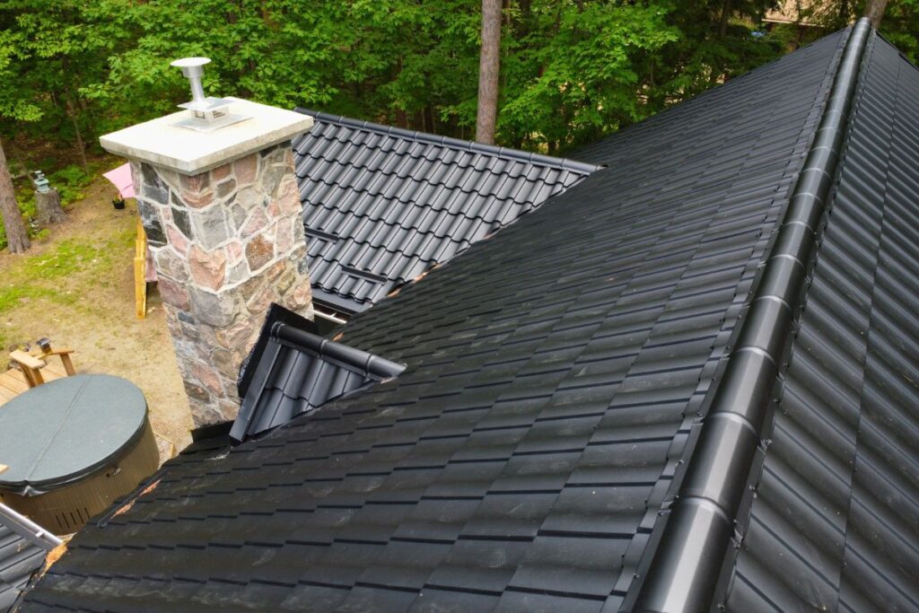 Close-up view of two black metal roof slopes intersecting, featuring a chimney and surrounded by lush greenery, demonstrating the roof's durability and neat construction.