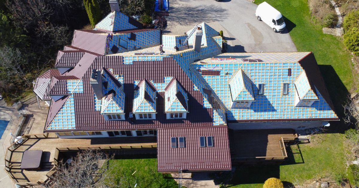 Complex multi-section residential roof with underlayment installed and partial metal roofing panels in progress, multiple dormers and valleys visible.