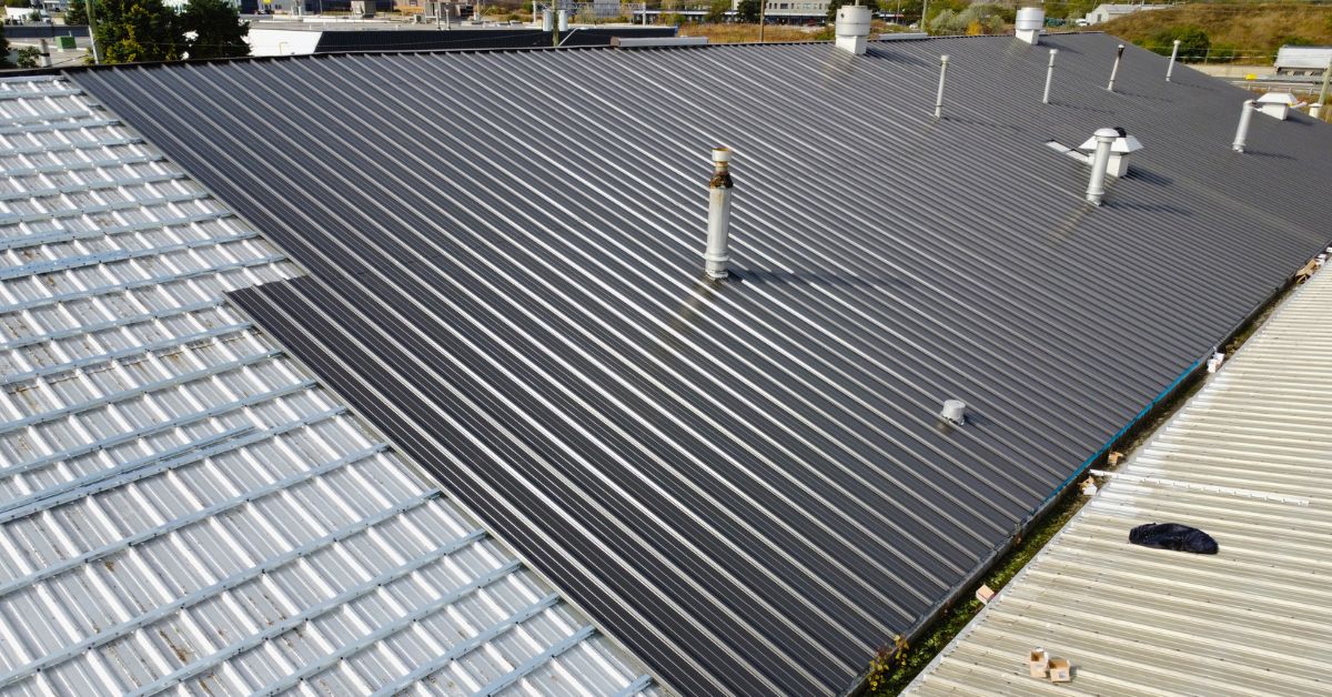 Commercial standing seam metal roof installed on an industrial building with rooftop ventilation pipes and adjacent corrugated roofing sections.